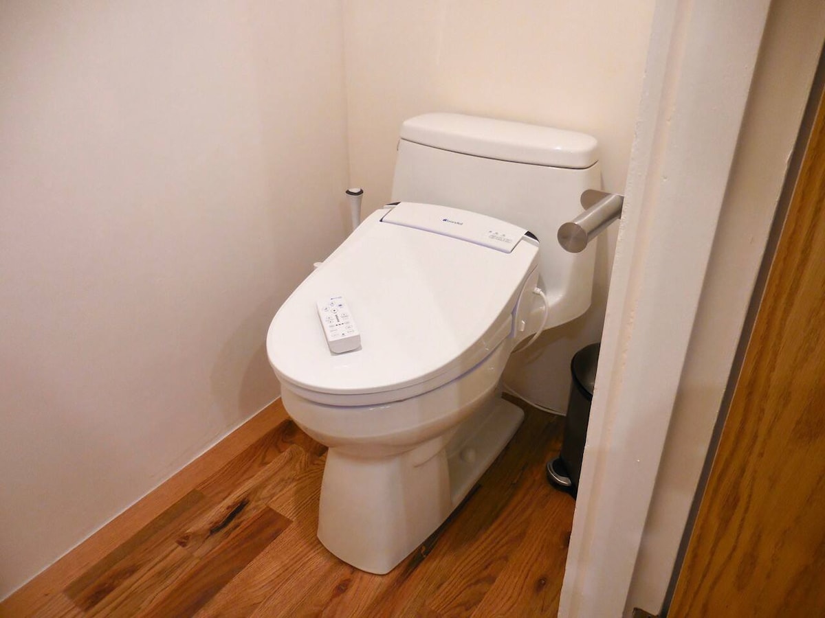 A modern toilet equipped with a heated bidet seat is positioned against a neutral wall. The sleek design is complemented by wooden flooring, enhancing the minimalist aesthetic of the space.