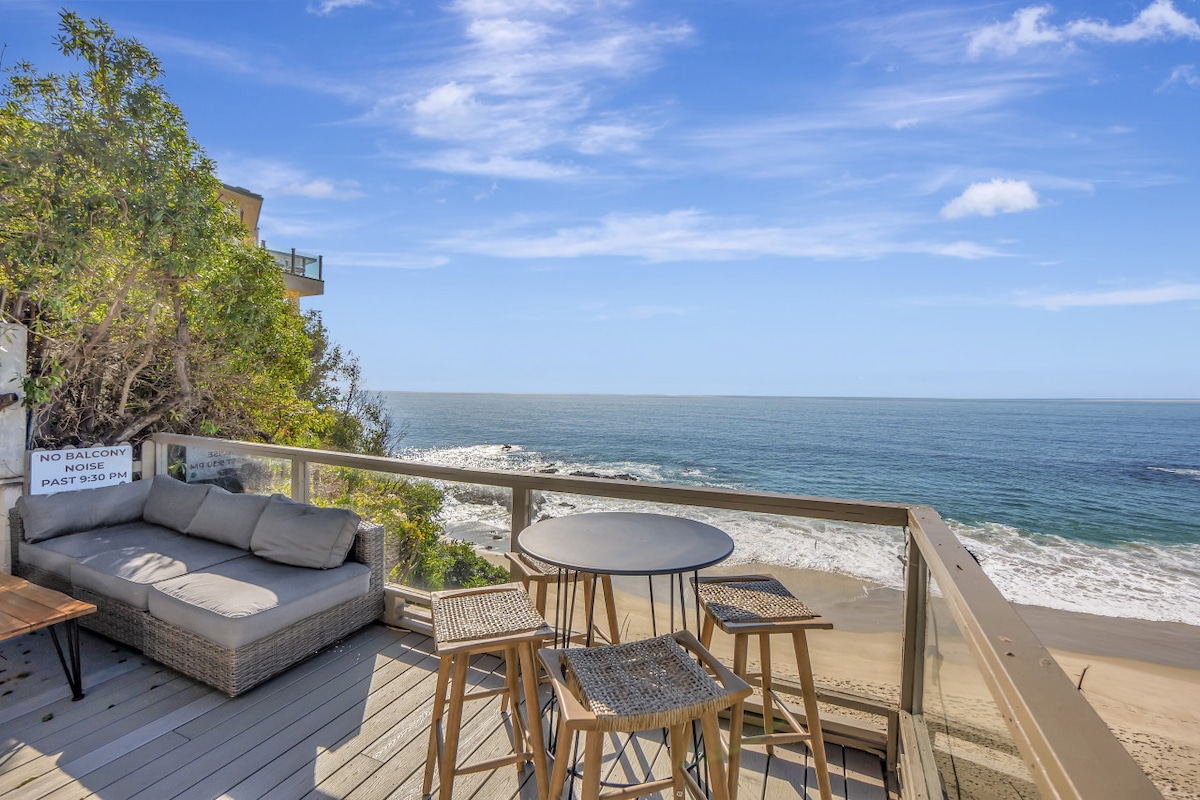 An outdoor deck is pictured, featuring a cozy seating area with a sectional sofa and a dining table surrounded by tall stools. The expansive ocean views and sandy beach are visible beyond the railing, with a clear blue sky overhead.