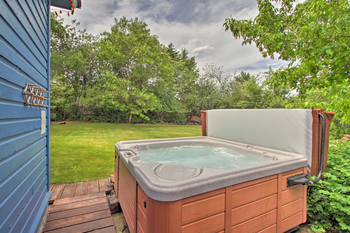 A private hot tub is positioned on a wooden deck, surrounded by lush greenery and a well-maintained grassy backyard. The inviting tub features a cover that is partially open, with gentle water movement visible on the surface, offering a relaxing outdoor setting.