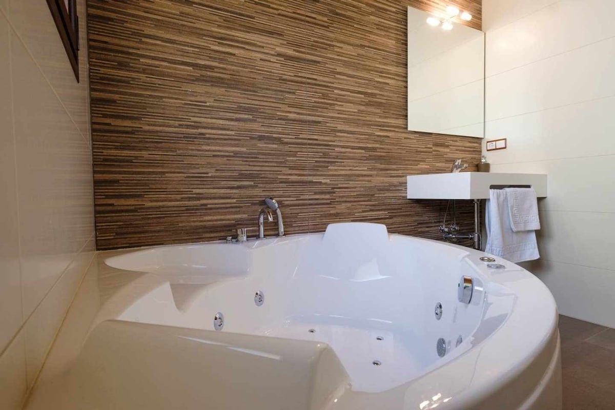 A spacious white jacuzzi is placed in a modern bathroom, complemented by a textured brown wall. A large mirror reflects light above the sink area, which includes neatly folded towels and a sleek countertop.