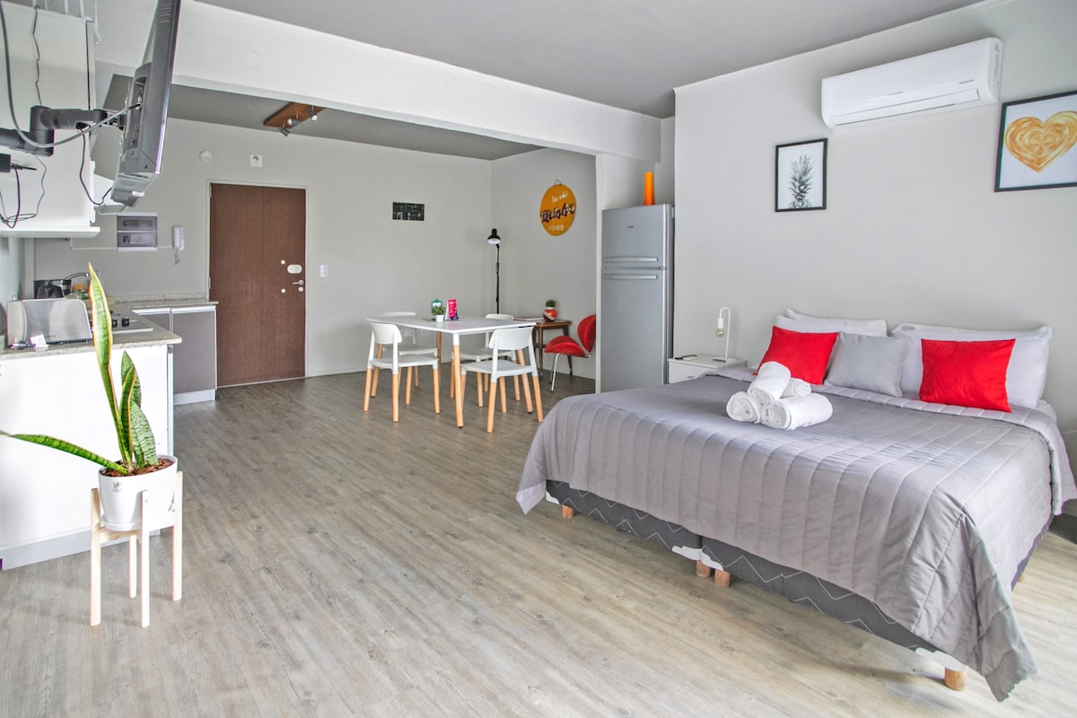 A spacious studio apartment features a generous sleeping area with a large bed, accented by red and gray textiles. The open layout includes a dining table for four, a sleek kitchen with a refrigerator, and light wood flooring throughout.