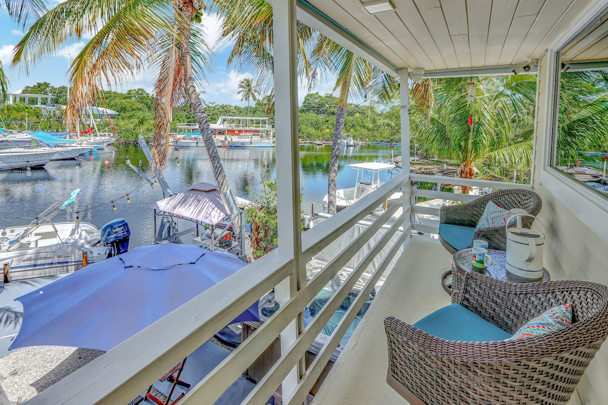 A covered balcony offers comfortable seating with two wicker chairs and a small side table. Lush palm trees frame the view of the tranquil waterfront, where boats are docked in calm waters. Sunlight filters through the space, creating a serene atmosphere.