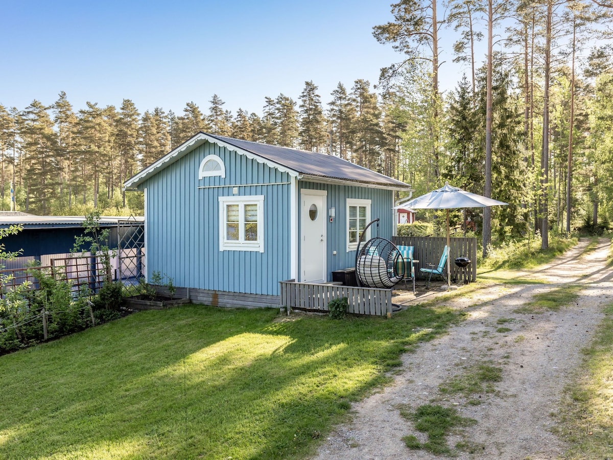 A small timber house with a blue exterior is surrounded by trees, situated on a gravel path. The entrance features a round window and a door with a circular design. A shaded terrace with outdoor seating is visible, offering a peaceful atmosphere.