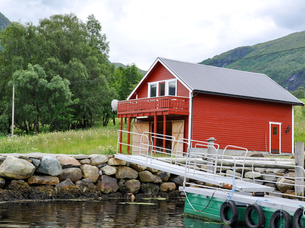 A bright red house is situated by the water's edge, with a wooden walkway leading to it. The structure features a balcony and large windows, allowing views of the surrounding landscape. Green hills and trees frame the scene, enhancing the natural setting.