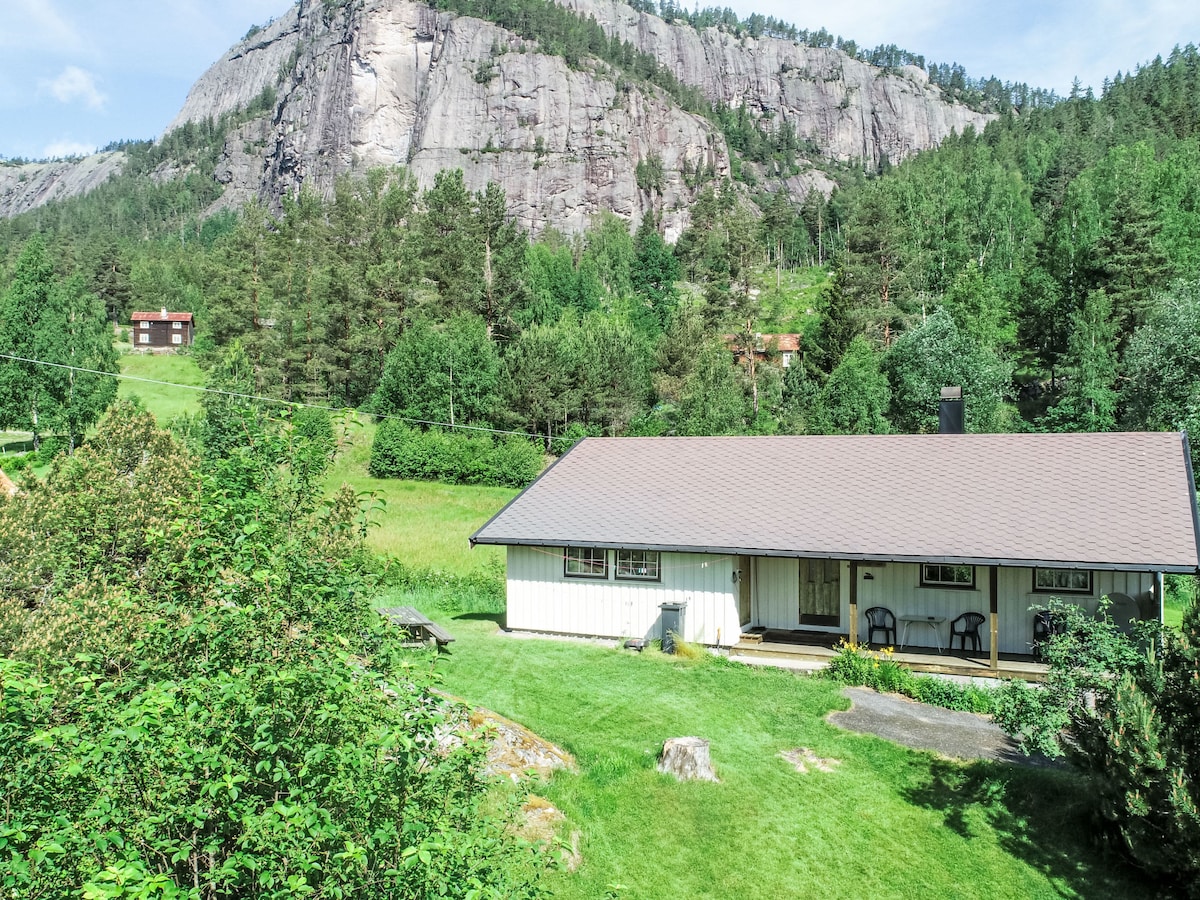 A single-story house is set amidst lush greenery, featuring a well-maintained lawn and a stone pathway leading to the entrance. The backdrop is formed by a towering rock face, surrounded by dense trees. Outdoor seating areas are visible, providing a serene setting.
