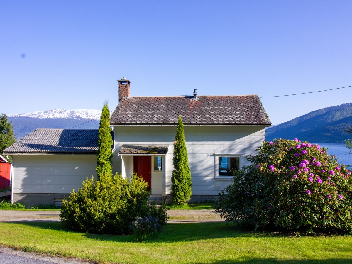 A charming two-level house is set against a clear blue sky, featuring a stone chimney and wooden roof. Lush green bushes and vibrant flowers surround the entrance, while a scenic view of mountains and water can be seen in the background.