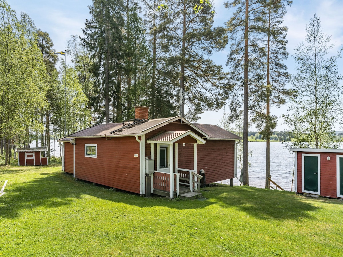 A red timber house is situated on a grassy area by the lake, surrounded by tall trees. The structure features a porch and windows that offer a view of the water. Additional outbuildings can be seen nearby, enhancing the natural setting.