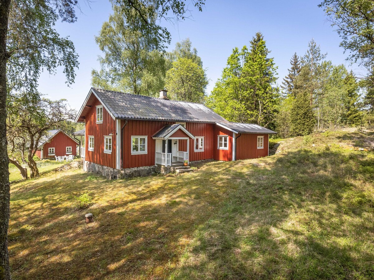 A charming red wooden house is surrounded by a vibrant natural landscape, featuring trees and grassy areas. The house showcases a welcoming entryway and is designed with multiple windows, allowing natural light to fill the interior. The peaceful countryside setting enhances the serene atmosphere.
