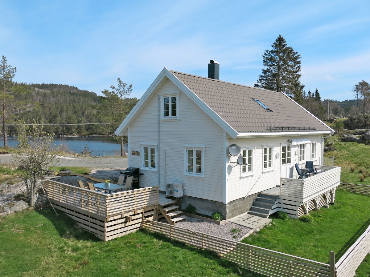 The exterior of a two-story house is presented, featuring a light-colored facade and a pitched roof. A spacious deck with outdoor furniture is visible in the foreground, alongside a lush green lawn. The serene lake is seen in the background, framed by trees.