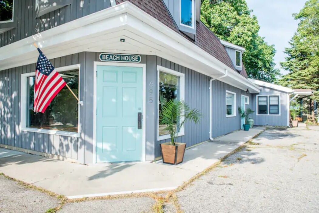 The exterior of the property features a light blue door marked with the sign 'BEACH HOUSE.' An American flag hangs beside the entrance. Large windows allow natural light to fill the space, and potted plants are positioned by the entryway. The surrounding area is paved.
