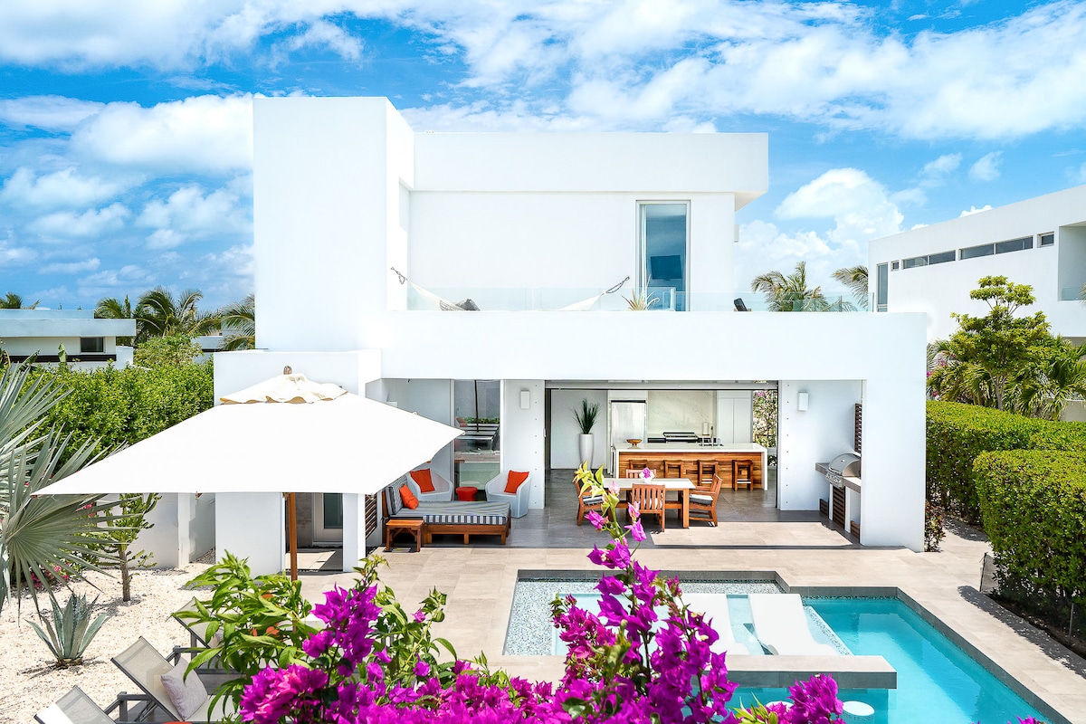 An exterior view of the villa showcases a modern design with clean lines and bright white walls. A pool is bordered by lush greenery and vibrant purple flowers. A shaded seating area complements the outdoor dining space, with large windows reflecting the blue sky.