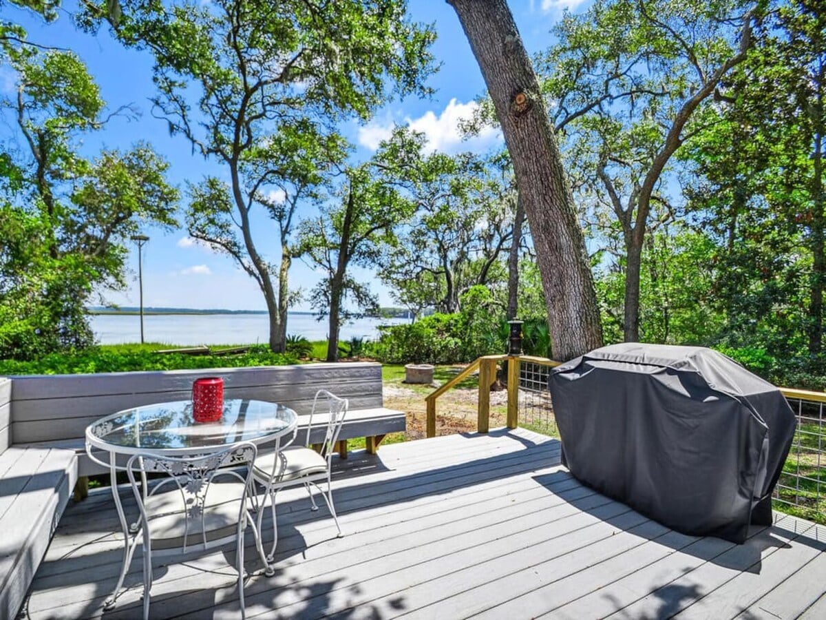 A spacious deck overlooks a serene landscape, featuring a round table with elegant seating and a covered grill. Surrounding trees provide shade, while the view of the river enhances the outdoor ambiance.