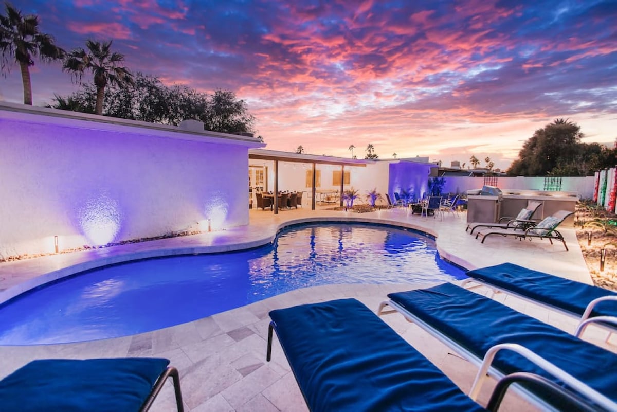 A serene pool area is framed by lounge chairs, featuring a blue pool with gentle curves. Soft purple and pink hues color the sky at sunset, reflecting off the water's surface. Palm trees stand in the background, enhancing the private outdoor setting.