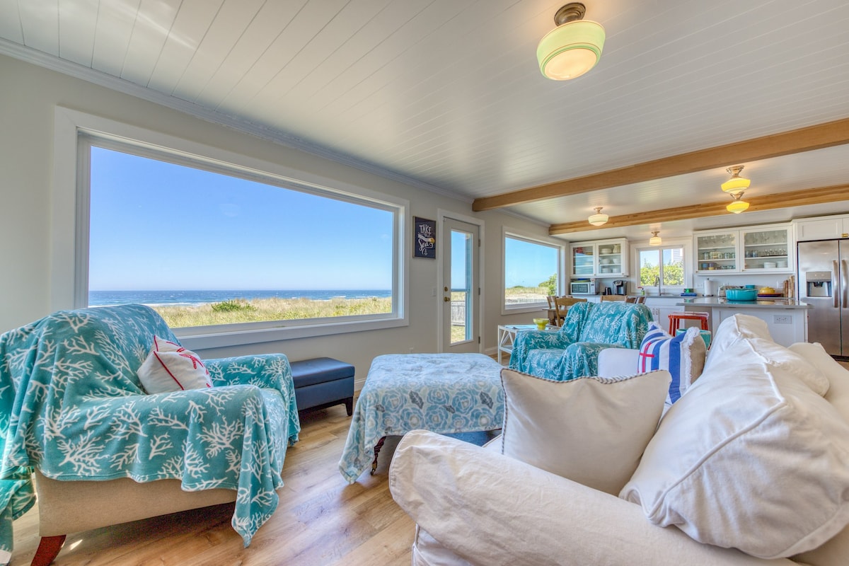 A bright living area features cozy seating arranged around a large window, showcasing expansive views of the ocean and beach. Light-colored furnishings are accented with colorful throws, while a fully equipped kitchen can be seen in the background, promoting an open and inviting atmosphere.