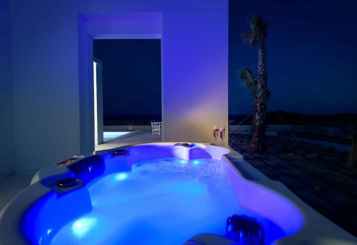 A hot tub is illuminated with soft blue lighting, situated on a terrace that offers a view of the sea. Glasses are placed on the edge, adding to the relaxing ambiance. The backdrop features an open doorway leading to another area, enhancing the serene atmosphere.