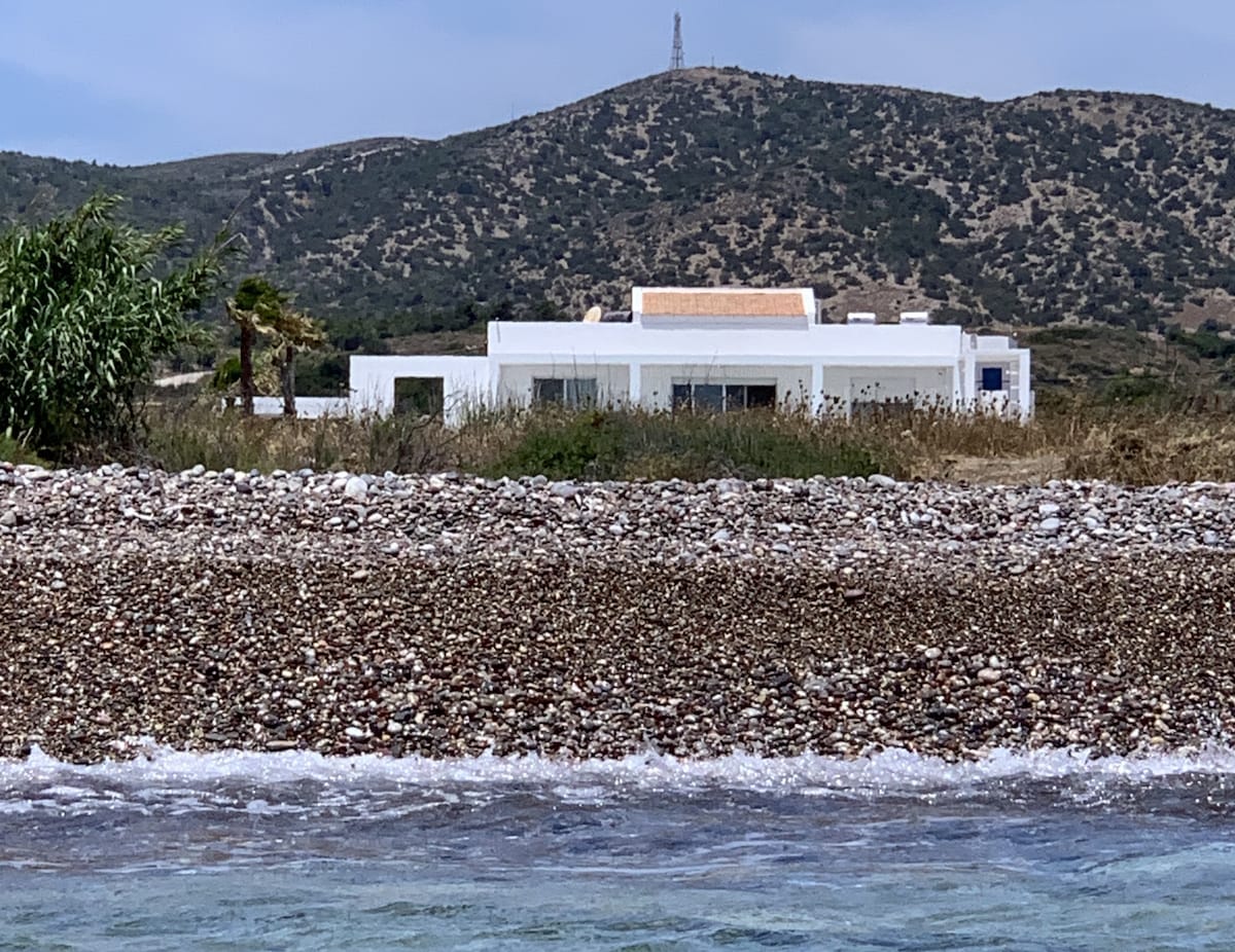 A modern beachfront villa is positioned against a backdrop of rolling hills. The structure, primarily white, features multiple windows reflecting the coastal scenery. The shoreline is made up of smooth pebbles, with gentle waves lapping at the edge, creating a serene atmosphere.