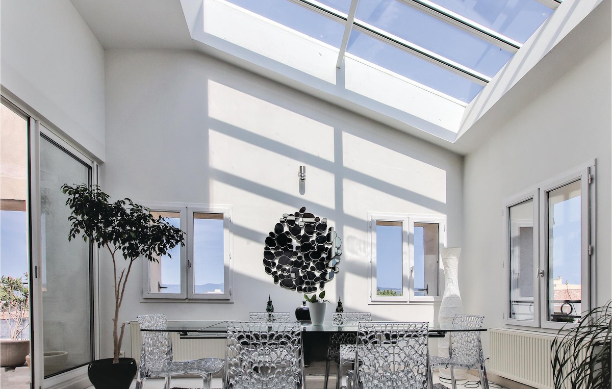 A bright dining area features a glass table surrounded by transparent chairs. A contemporary art piece adds visual interest to the wall. Natural light pours in through the skylights, creating an airy atmosphere and highlighting the sleek design of the space.