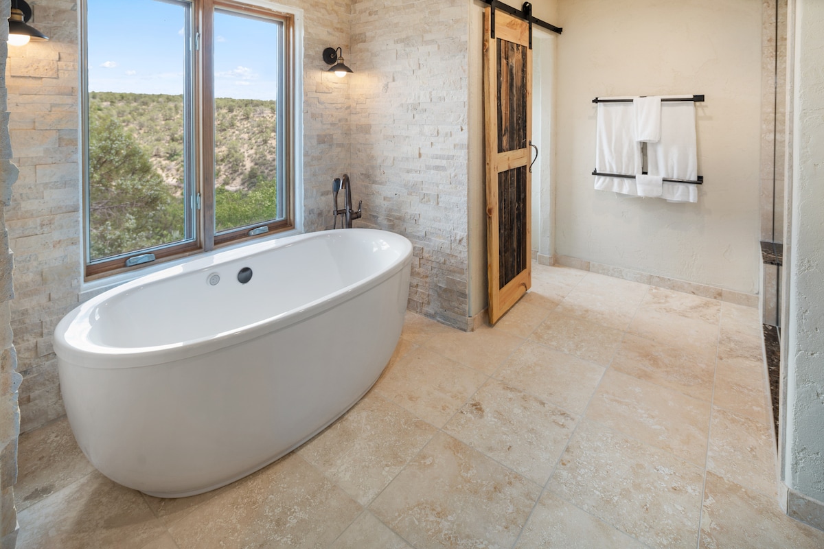 A spacious bathroom features a modern freestanding bathtub positioned near large windows that offer scenic views of the surrounding landscape. The natural stone walls and tile flooring create a serene ambiance. A wooden barn door adds a rustic touch, alongside neatly arranged towels on a rack.