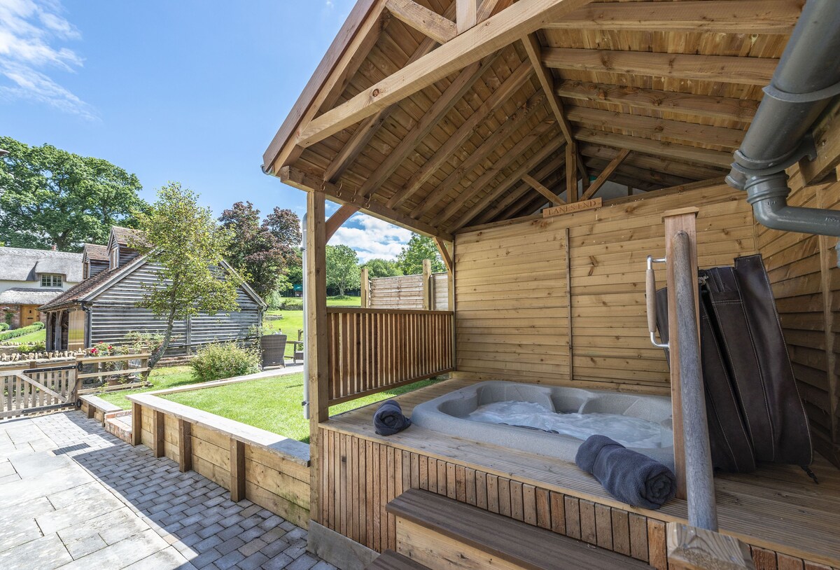 An outdoor hot tub is nestled under a wooden shelter, surrounded by a fenced area and garden. Soft towels are placed on the edge of the tub. A well-maintained lawn and colorful flower beds can be seen in the distance, along with charming cottages.