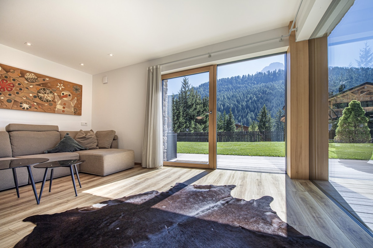 A spacious living area features a comfortable sofa and two modern tables. Large glass doors invite natural light and offer views of the lush green garden and surrounding mountains. A soft area rug adds warmth to the wood floor, enhancing the overall ambiance.