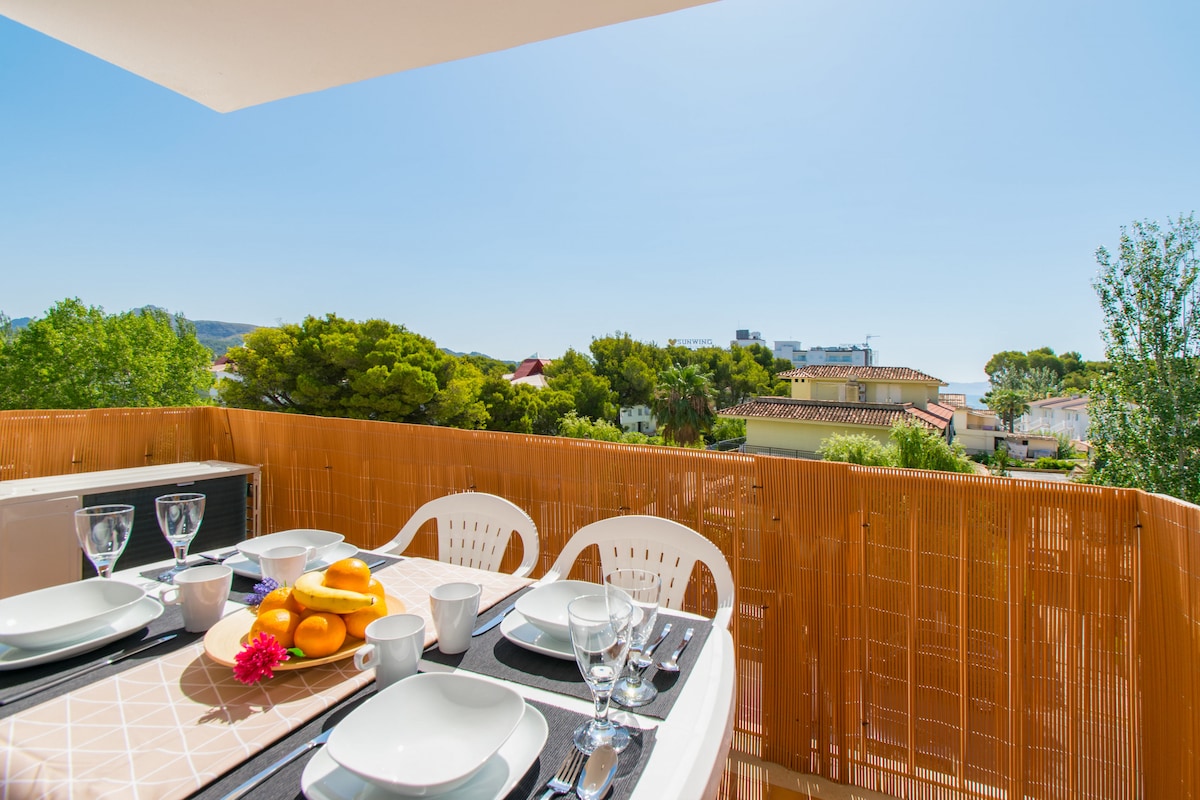A sunny terrace is shown, featuring a dining setup with a table neatly arranged with plates, glasses, and a centerpiece of fruit. The vibrant greenery of nearby trees is visible, alongside a clear blue sky, enhancing the inviting outdoor dining experience.