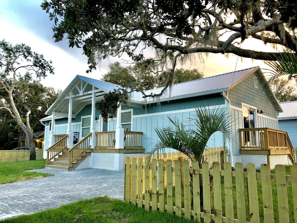 The exterior of a newer beach home is depicted, featuring a light blue facade and a welcoming porch with a gabled roof. The property is enclosed by a low wooden fence and surrounded by grassy areas and mature trees, providing shade and a relaxed atmosphere.