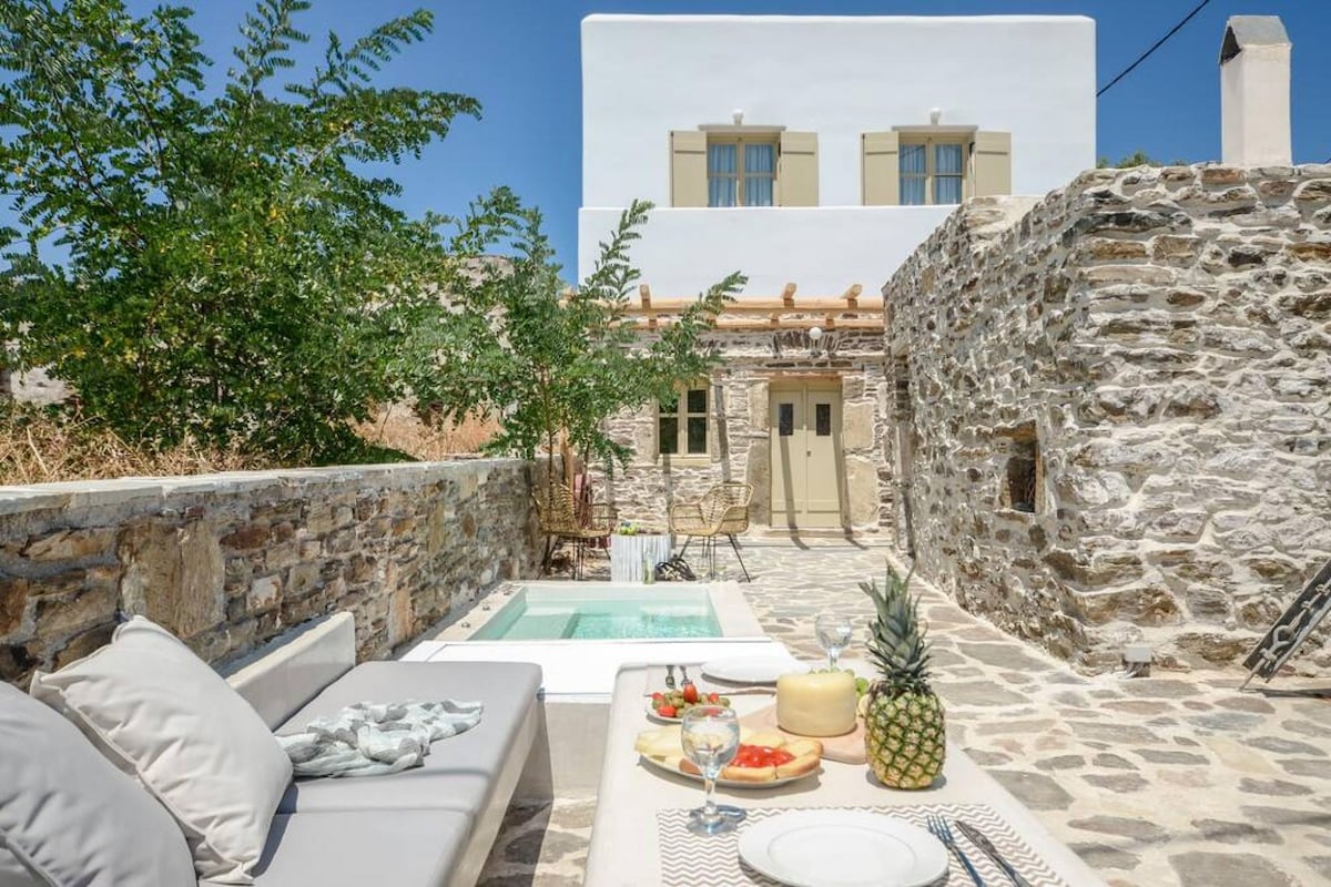A stone-paved outdoor area features a small hydromassage tub surrounded by lush greenery. A dining table is set elegantly with glasses and fruit, complementing the inviting seating area. The traditional stone walls enhance the rustic charm of this serene space.