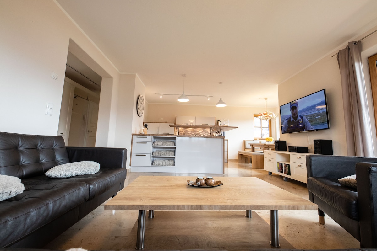 A spacious living area is visible, featuring two dark leather sofas arranged around a light wooden coffee table. A flat-screen TV is mounted on the wall, while an open kitchen with modern appliances and a dining space can be seen in the background.