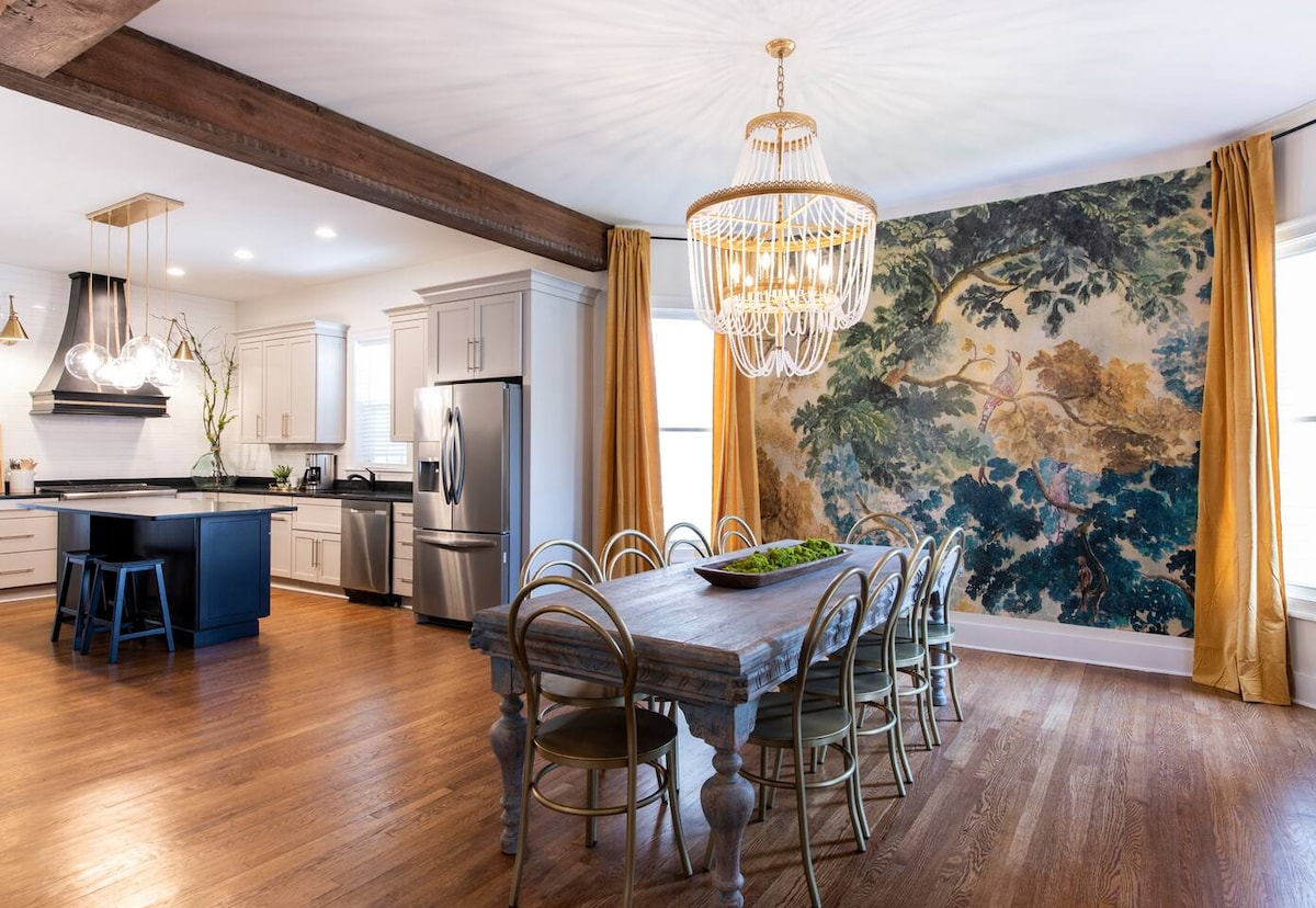 A modern kitchen and dining area combine functionality with style. The dining table, surrounded by elegant chairs, is illuminated by a chandelier. A large mural featuring nature scenes adorns one wall, contrasting against the soft-toned cabinetry and spacious island.