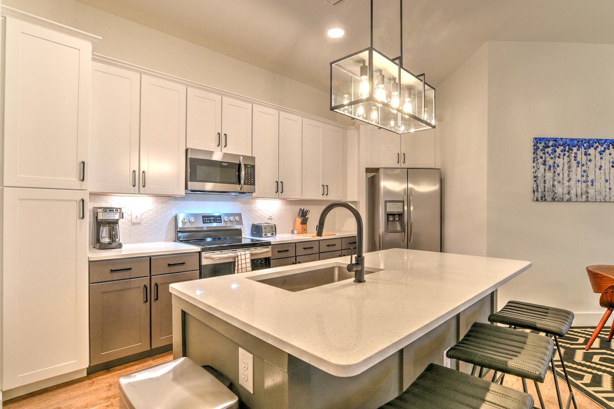 A modern kitchen is presented, featuring a central island with bar seating and sleek black stools. Stainless steel appliances, including a refrigerator and oven, complement the light cabinetry. Under-cabinet lighting illuminates the countertop, enhancing the clean lines and contemporary design.