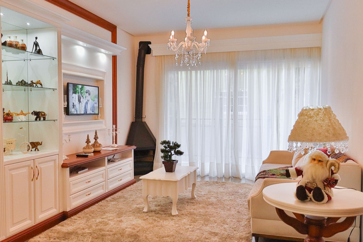 A well-lit living area features a modern television mounted above an elegant console. A plush area rug covers the floor, while a chandelier hangs from the ceiling. Large curtains allow natural light to fill the space, complemented by a cozy sofa and decorative accessories.