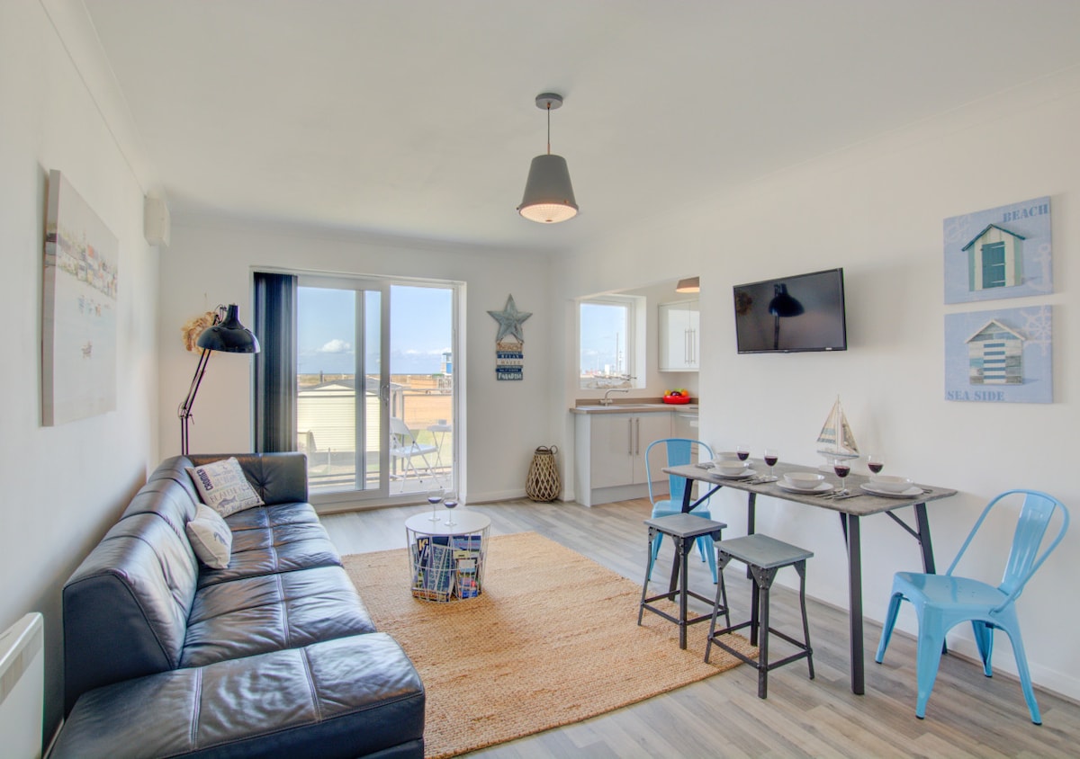 A spacious living area is presented with a dark leather sofa and a light rug. A wooden dining table with blue chairs is positioned near a window, allowing for natural light. Wall art depicts coastal themes, and a television is mounted on one wall.