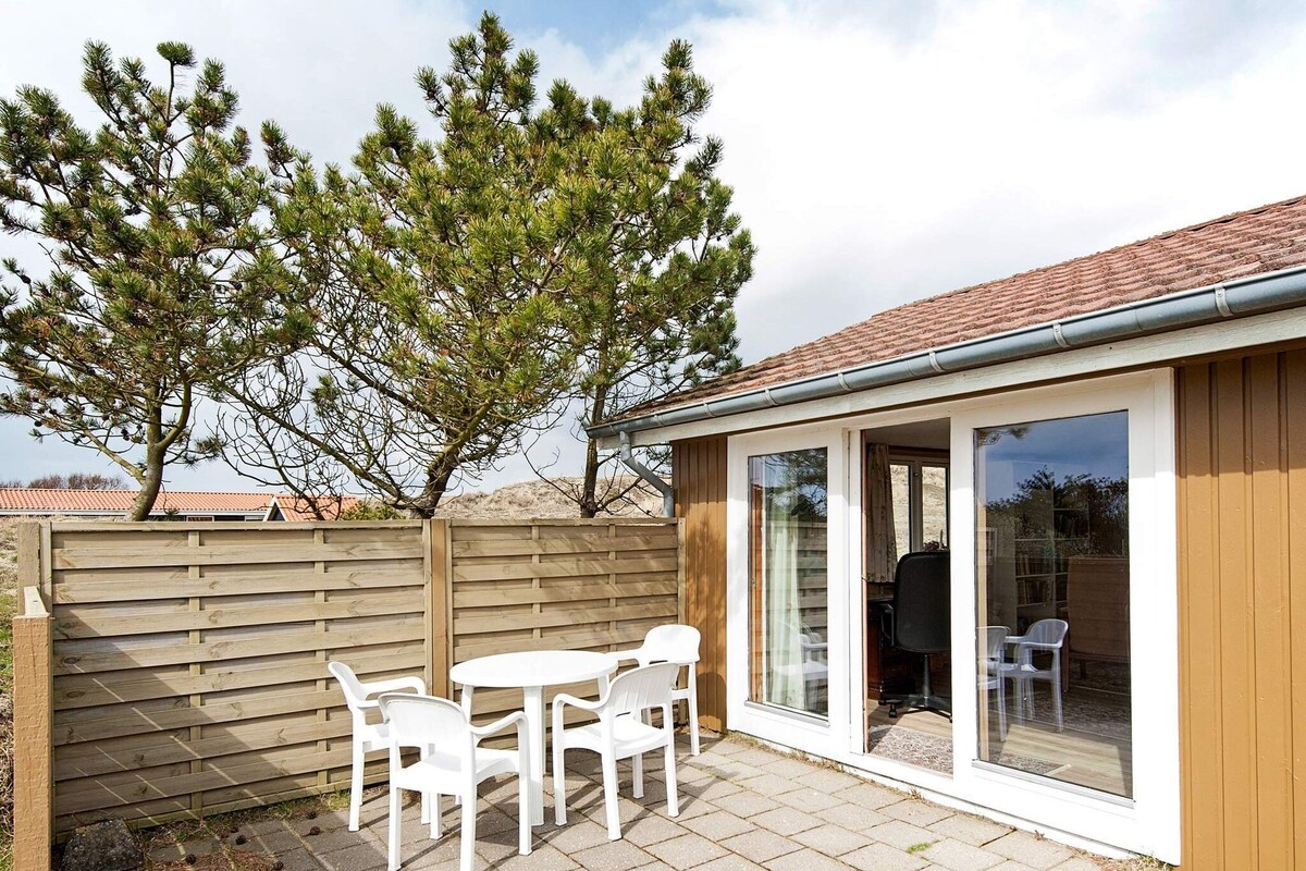 A west-facing terrace is equipped with a round table and four white chairs, set on stone paving. The space is surrounded by a wooden fence and tall trees, offering a peaceful atmosphere. Large glass doors provide access to the interior of the holiday home.