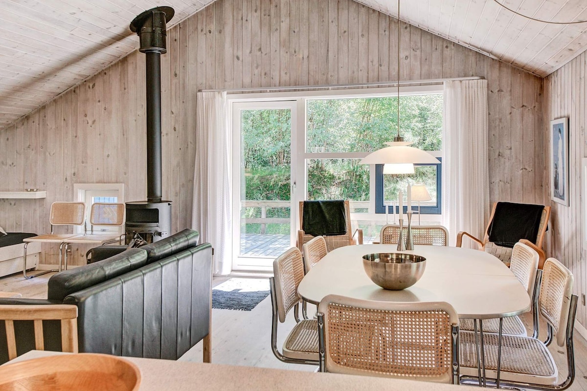 A spacious living area features wooden walls and a ceiling with a light finish. A large window offers natural light, with views of greenery outside. A modern dining table is surrounded by chairs, and a cozy seating arrangement includes an armchair and a fireplace.