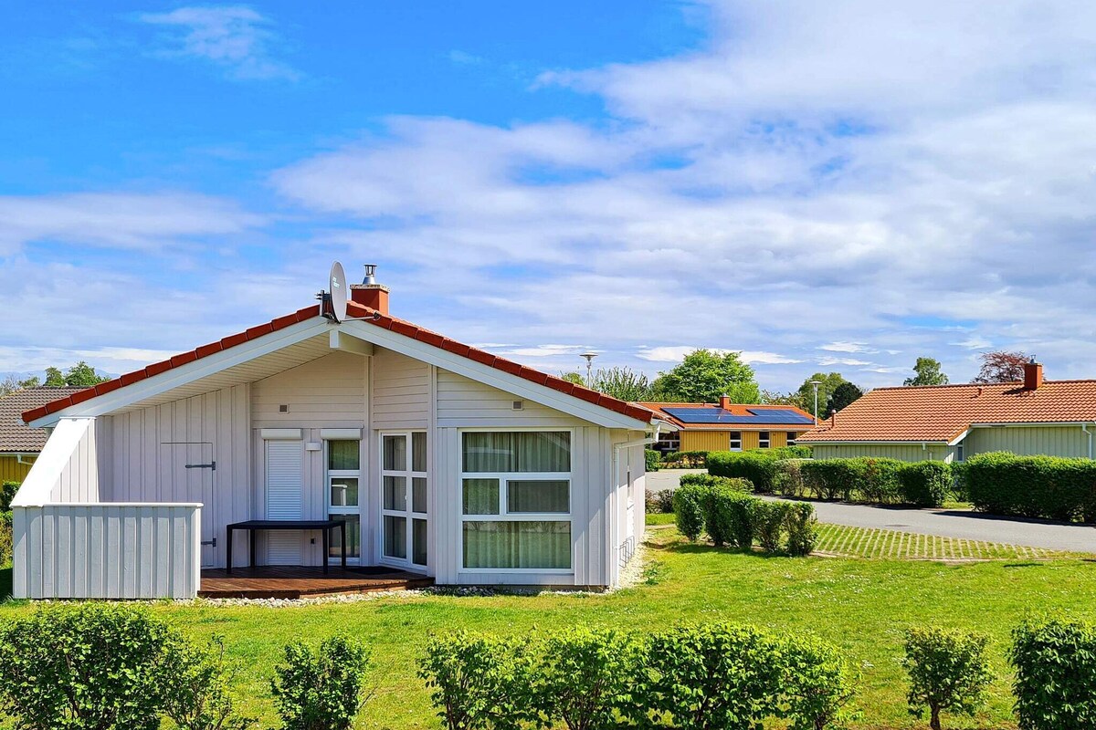 A single-story cottage is shown, featuring a sloped roof and large windows that provide ample natural light. The exterior is mostly white, complemented by a wooden terrace. Lush green lawns and neatly trimmed hedges surround the property, with other homes visible in the background.