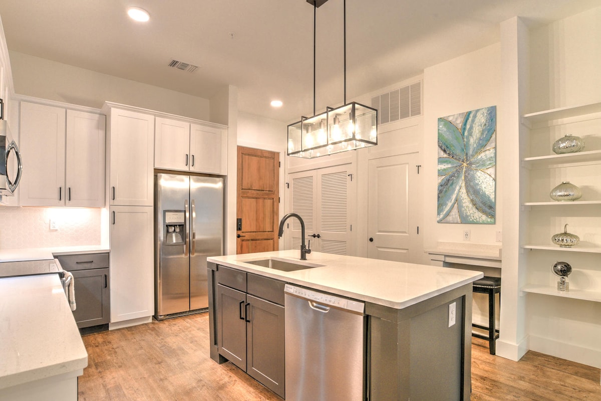 An open kitchen is highlighted by modern stainless steel appliances, including a refrigerator and dishwasher. A large island serves as a functional workspace, and a contemporary light fixture hangs above. Neutral cabinetry complements the light-colored walls, contributing to an airy and well-lit environment.