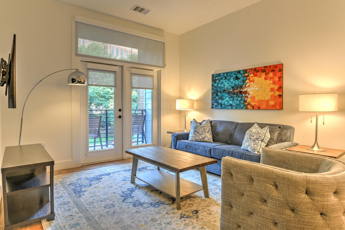 A well-furnished living area features a plush sofa, a coffee table positioned in the center, and two lamps providing soft illumination. A large wall art piece adds color to the space, while double doors lead to an outdoor area, enhancing natural light.