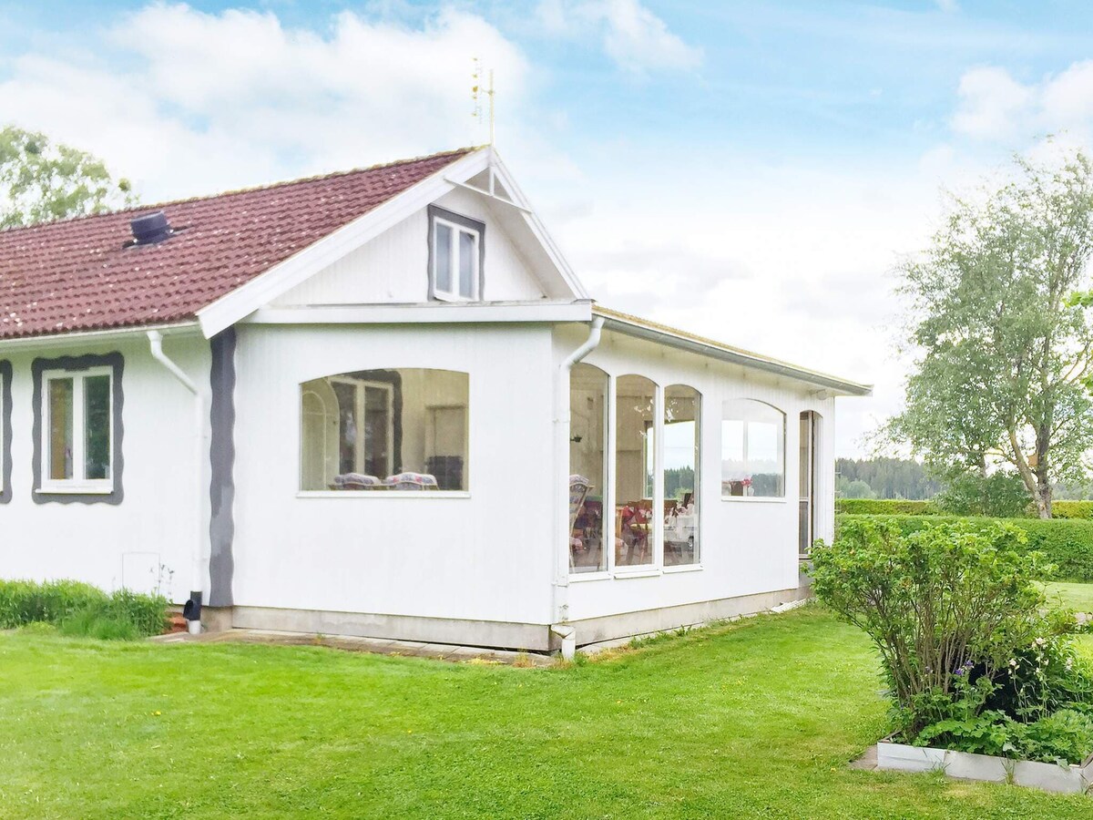 The exterior of the holiday home is shown, featuring a white facade with a brown tiled roof. The conservatory with large glass windows extends outward, offering views of the surrounding green lawn and flatlands. Lush greenery and shrubs are visible in the garden area.