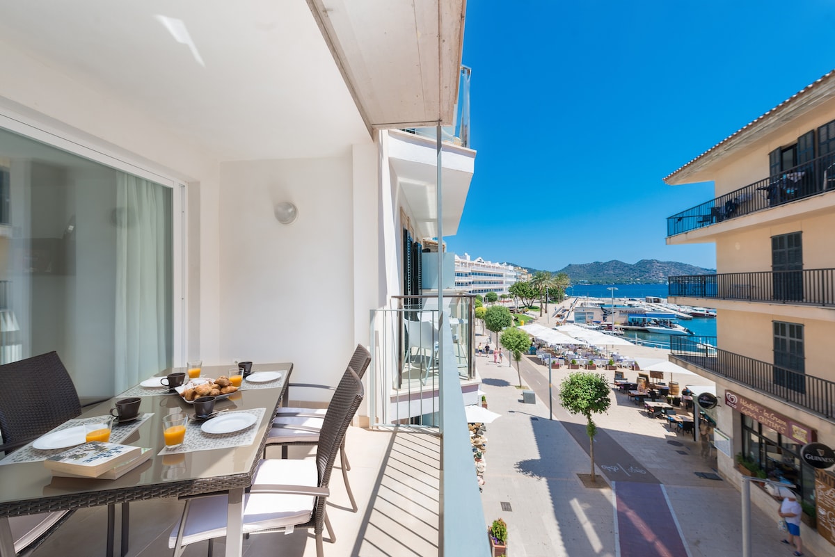 A terrace is showcased with a dining table set for breakfast, including plates and beverages. The view features a clear blue sky, the marina with boats, and the surrounding coastal landscape, providing a scenic backdrop for outdoor dining.