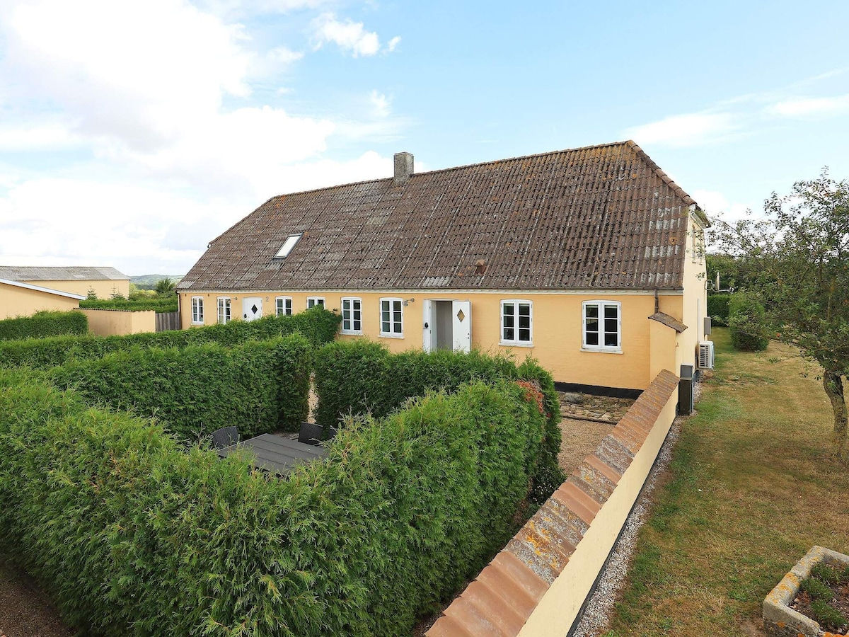 The exterior of a two-story holiday home is surrounded by lush hedges, providing privacy. The sloping roof and yellow facade complement the natural landscape. A terrace area is visible in front, creating a welcoming space for outdoor relaxation.