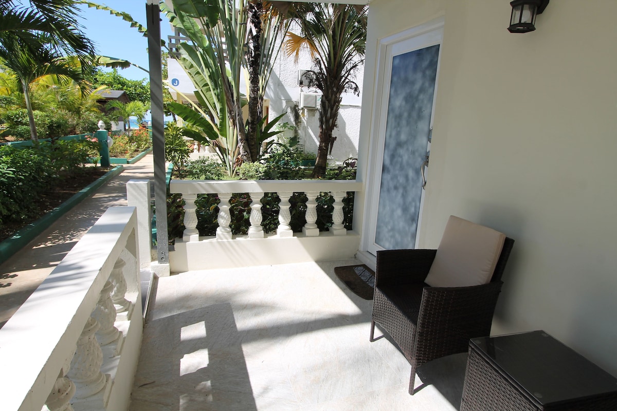 A furnished patio area offers a comfortable chair and a small table, surrounded by tropical plants. The doorway leads into the suite, providing easy outdoor access. Sunlight filters through palm leaves, creating a serene outdoor atmosphere.
