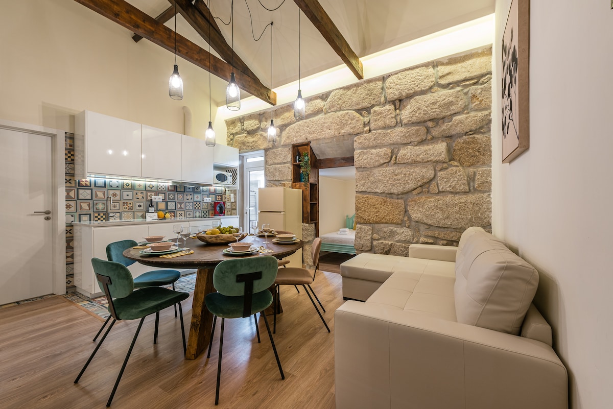 An open dining area features a round wooden table set for six, surrounded by green chairs. A spacious sofa is positioned nearby, and rustic stone walls enhance the setting. Modern kitchen cabinetry is visible in the background, complemented by stylish lighting fixtures.