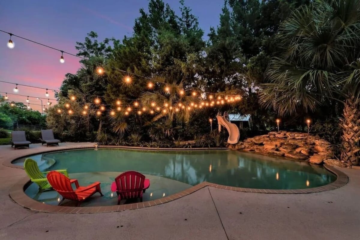 A resort-style pool is surrounded by lush greenery and features a waterslide. Colorful lounge chairs are positioned nearby, and string lights create a warm ambiance overhead as the sky transitions into twilight.