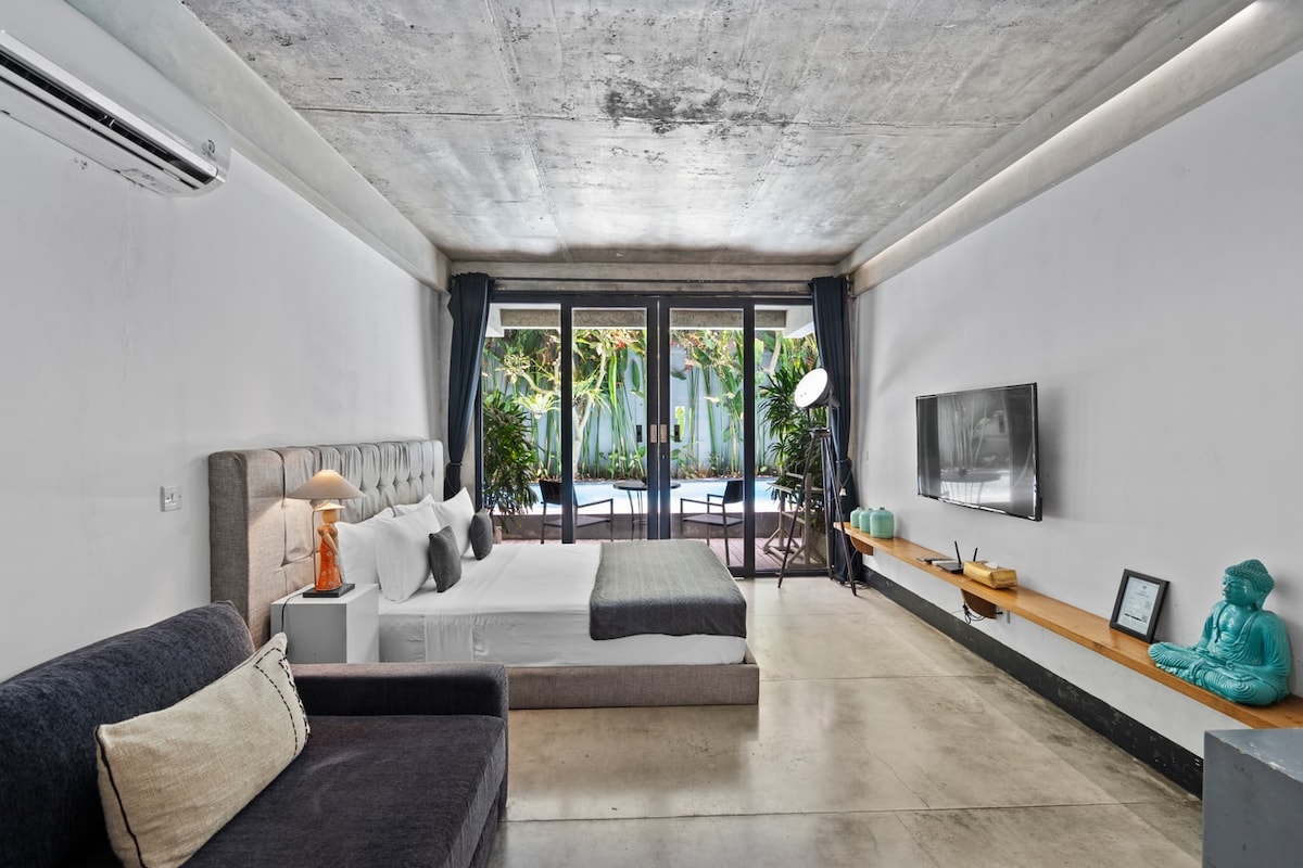 A spacious bedroom features a king-size bed dressed in neutral linens, complemented by artistic wall decor. A cozy sofa is present, with floor-to-ceiling glass doors providing views of lush greenery and direct access to the outdoor terrace. Minimalist furnishings enhance the modern aesthetic.