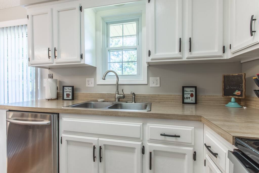 A modern kitchen features white cabinetry and a stainless steel sink with dual basins. Light wooden countertops provide workspace, and a window above the sink allows natural light to enter. Cooking essentials and decorative items are neatly arranged on the countertops.