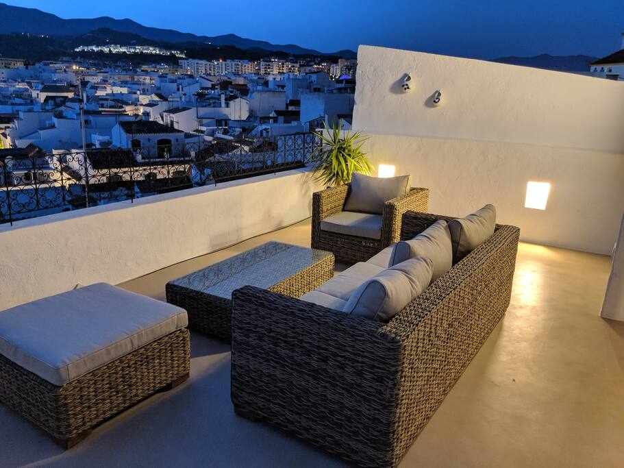 An inviting rooftop terrace features comfortable wicker seating arranged around a low table. Soft cushions enhance relaxation, while the surrounding walls provide a sense of privacy. The view of the town below extends to the mountains in the distance under a twilight sky.