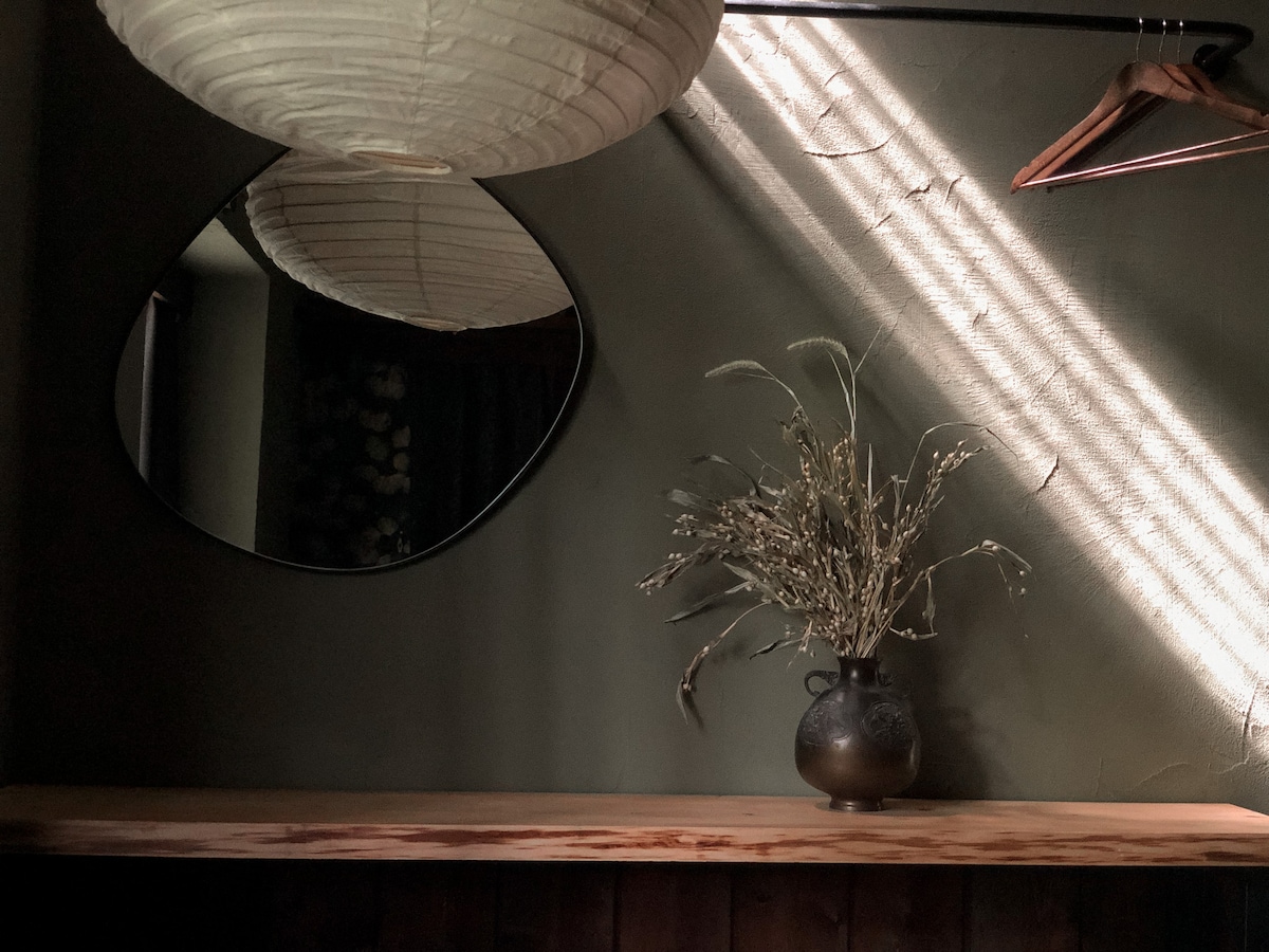 A softly lit corner features a round mirror reflecting the warm light from a hanging pendant. A wooden shelf holds a dark vase with dried flowers, while gentle shadows play across the textured green wall.