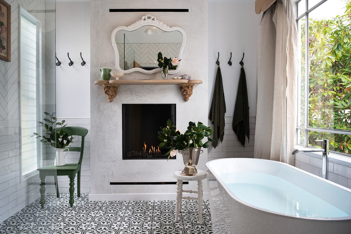 An elegant bathroom features a freestanding tub positioned beside a charming gas log fireplace. Decorative tiles with intricate patterns cover the floor. Natural light streams in from a large window, highlighting greenery visible outside, while a mirror above a wooden shelf adds a touch of sophistication.