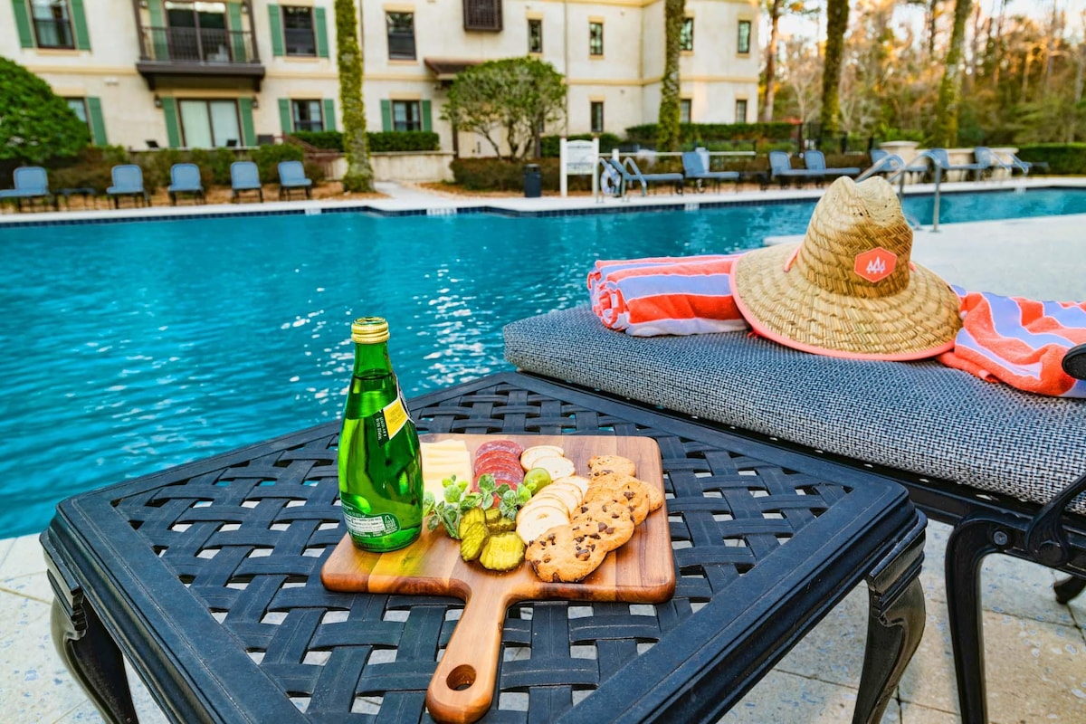 A wooden serving board displays an assortment of snacks, including cookies and sliced vegetables, positioned on a textured black table. A green sparkling water bottle sits beside the board. A striped towel and a straw hat are neatly arranged on a nearby lounge chair, with a sparkling pool in the background.