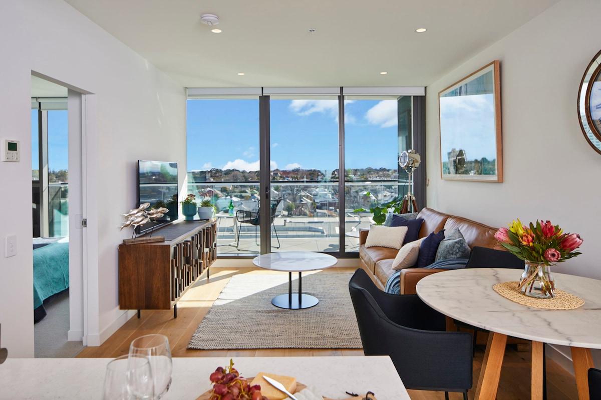 A well-appointed living space showcases large glass doors providing access to a balcony with expansive views. A plush leather sofa is positioned alongside a marble coffee table. A round dining table comfortably seats four. Bright natural light fills the area, highlighting elegant furnishings and decor.